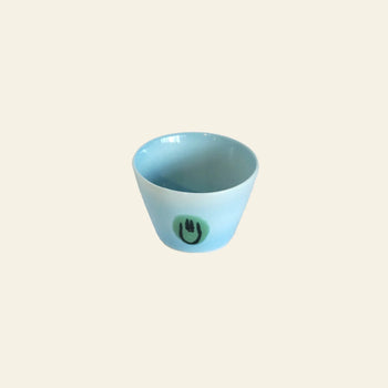 The Frizbee Ceramics Super Cup L by Frizbee Ceramics is a small, light blue ceramic cup with a glossy finish. It features a simple green circle and black smiley face—an artisanal, charming latte companion set against a beige background.