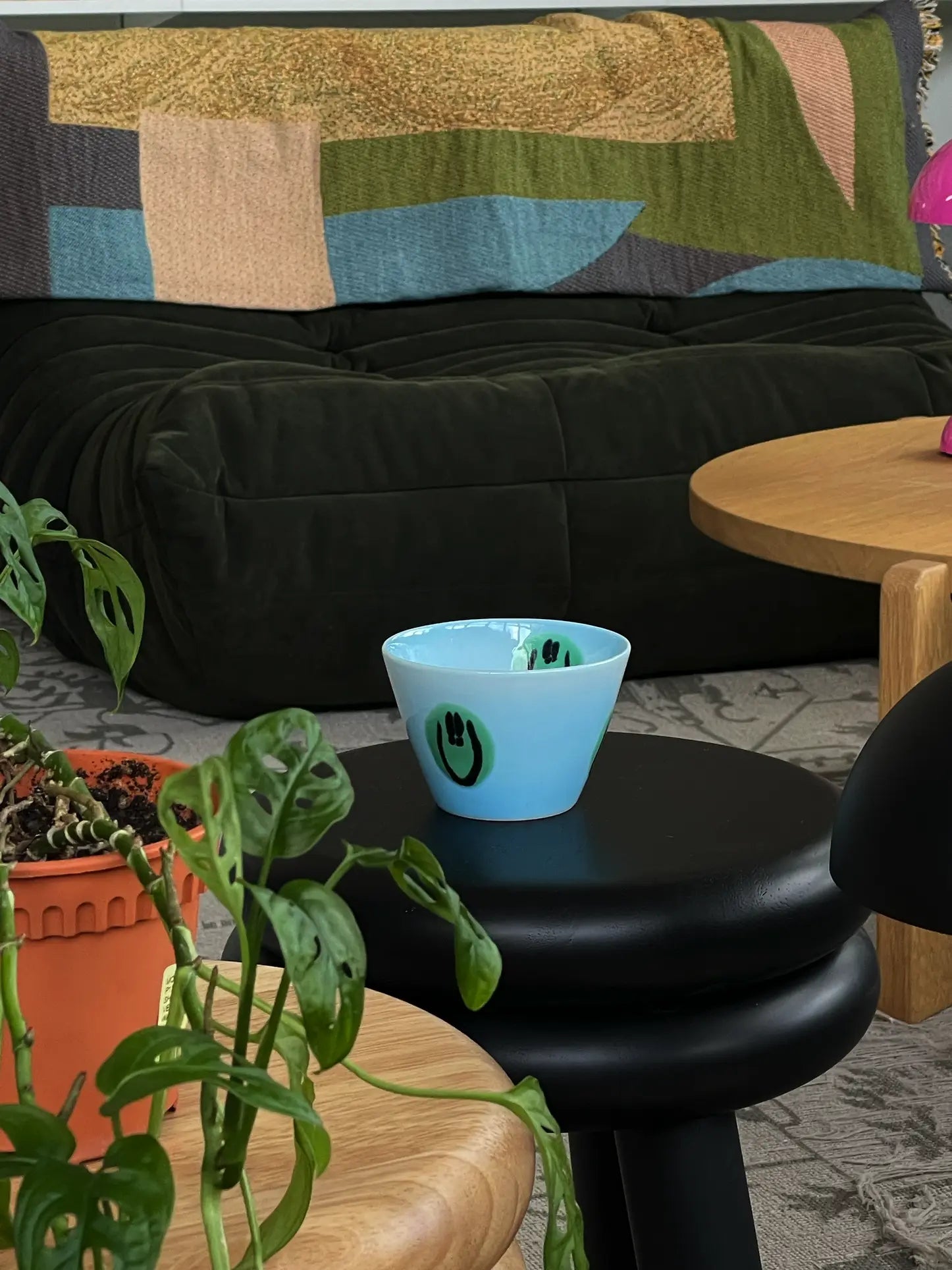 The Frizbee Ceramics Super Cup L by Frizbee Ceramics, in light blue with green and black eye-like designs, rests on a black stool amid wooden tables, a leafy green plant, and a dark couch with a vibrant patterned blanket.