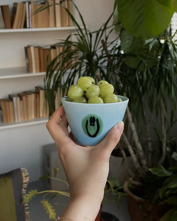 A hand holds the Frizbee Ceramics Super Cup L by Frizbee Ceramics, featuring a smiley face design and filled with green grapes. Its artisanal style pops against bookshelves, vibrant indoor plants, and soft window light.
