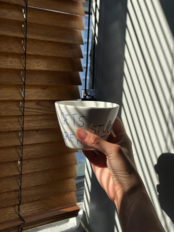 A hand holds the Frizbee Ceramics Super Cup L, a white ceramic bowl with blue script, by a sunlit window with wooden blinds—an artisanal piece from Frizbee Ceramics and an ideal companion for your latte.