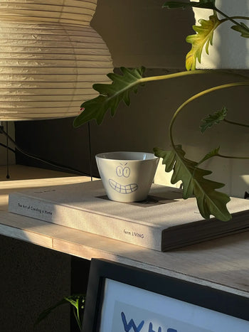 The Frizbee Ceramics Super Cup L, a charming white bowl with a grinning face by Frizbee Ceramics, rests on The Art of Creating a Home, surrounded by sunlight and green leaves for a cozy, inviting scene.