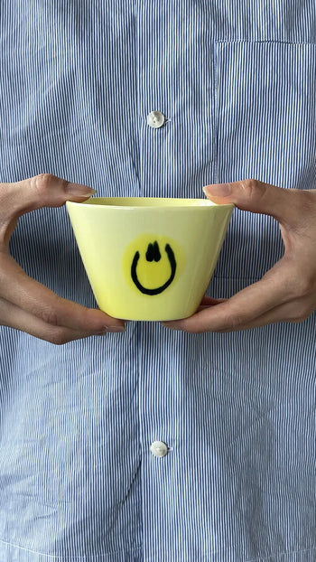 A person holds the Frizbee Ceramics Super Cup L, a yellow cup with a black smiley face, radiating artisanal charm—an ideal latte companion from Frizbee Ceramics.
