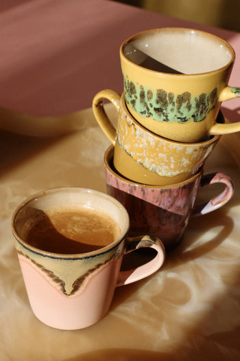 The HKLIVING 70s Ceramics: Americano Mugs, Set of 4, feature vibrant patterns and colors inspired by 70s ceramics; three are stacked and one pink mug is filled with coffee on a light marbled surface under warm lighting.