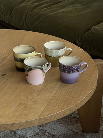 The HKLIVING 70s Ceramics: Americano Mugs, Set of 4 features hand-glazed stoneware in pastel shades and unique patterns, arranged on a round coffee table beside a green couch for retro charm.