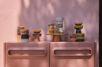 A set of HKLIVING 70s Ceramics Americano Mugs sits artfully on a pink cabinet, bathed in dappled sunlight and tree shadows across a pastel pink background.
