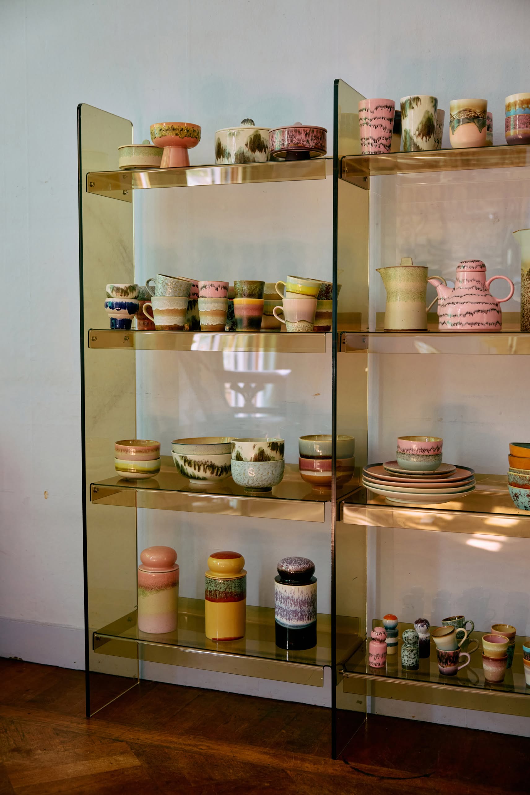 A glass shelving unit showcases HKLIVING 70s Ceramics: Americano Mugs, Set of 4, along with other pastel and earthy-toned ceramic cups, bowls, plates, and pitchers featuring unique patterns against a light-colored wall.
