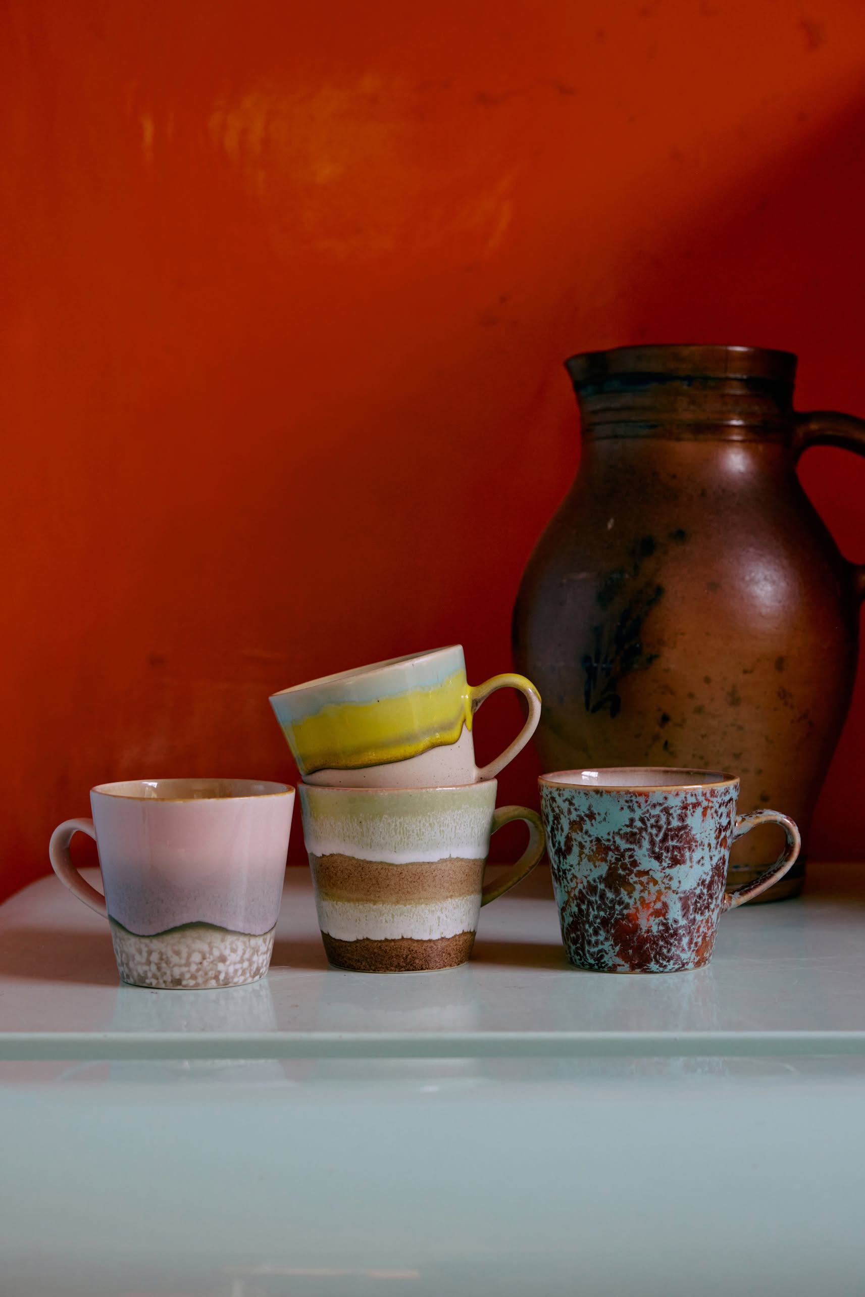 Four uniquely patterned HKLIVING 70s Ceramics: Americano Mugs (Set of 4) are stacked on a white surface, with a brown ceramic jug behind them against an orange backdrop, capturing the retro style characteristic of HKLIVING.
