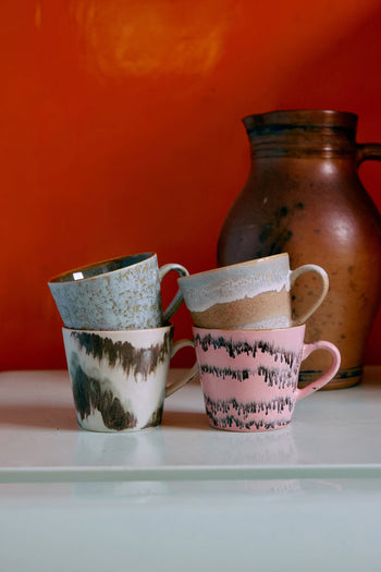 Four hand-glazed HKLIVING 70s Ceramics cappuccino mugs, each with a unique pattern, are stacked on a white surface before a large brown jug against an orange wall, capturing the perfect retro vibe.