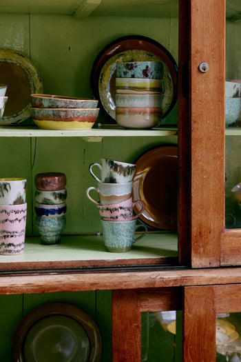 HKLIVING 70s Ceramics Cappuccino Mugs, Set of 4, are displayed with other painted cups, bowls, and plates inside a wooden glass-front cabinet with a green interior. Stacked mugs and hand-glazed patterns create an earthy retro look.
