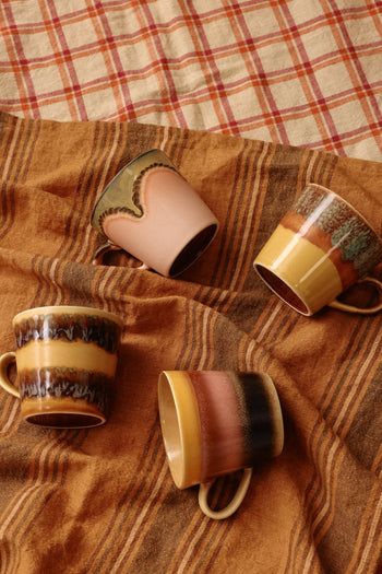 The HKLIVING 70s Ceramics Cappuccino Mugs, Set of 4, display earthy vintage designs in brown, tan, green, and yellow. They're arranged on a brown striped cloth with a red and cream plaid fabric in the background.