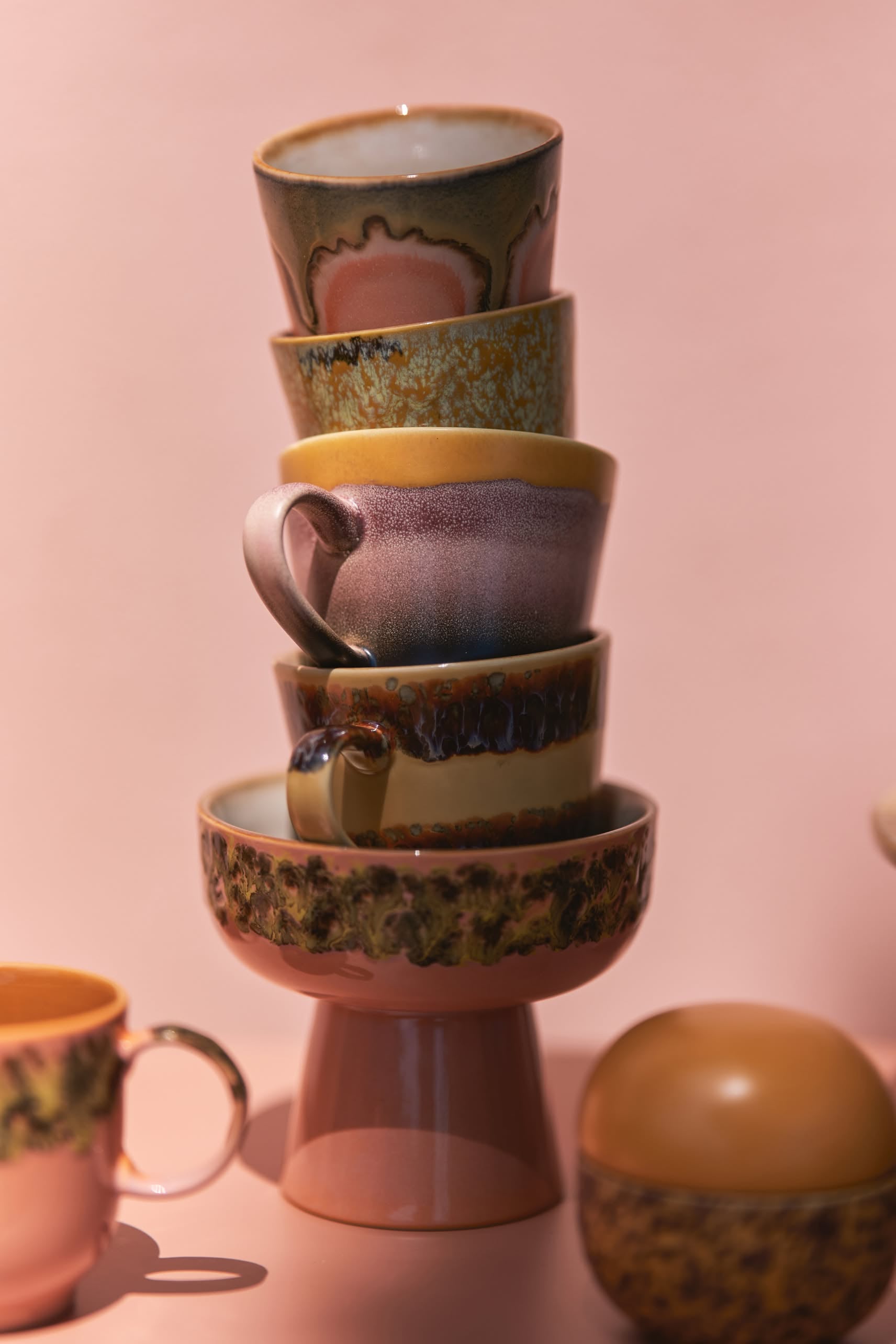 A stack of five hand-glazed ceramic cups and bowls in 70s patterns sits on a pink surface, with nearby HKLIVING 70s Ceramics: Cappuccino Mugs, Set of 4.
