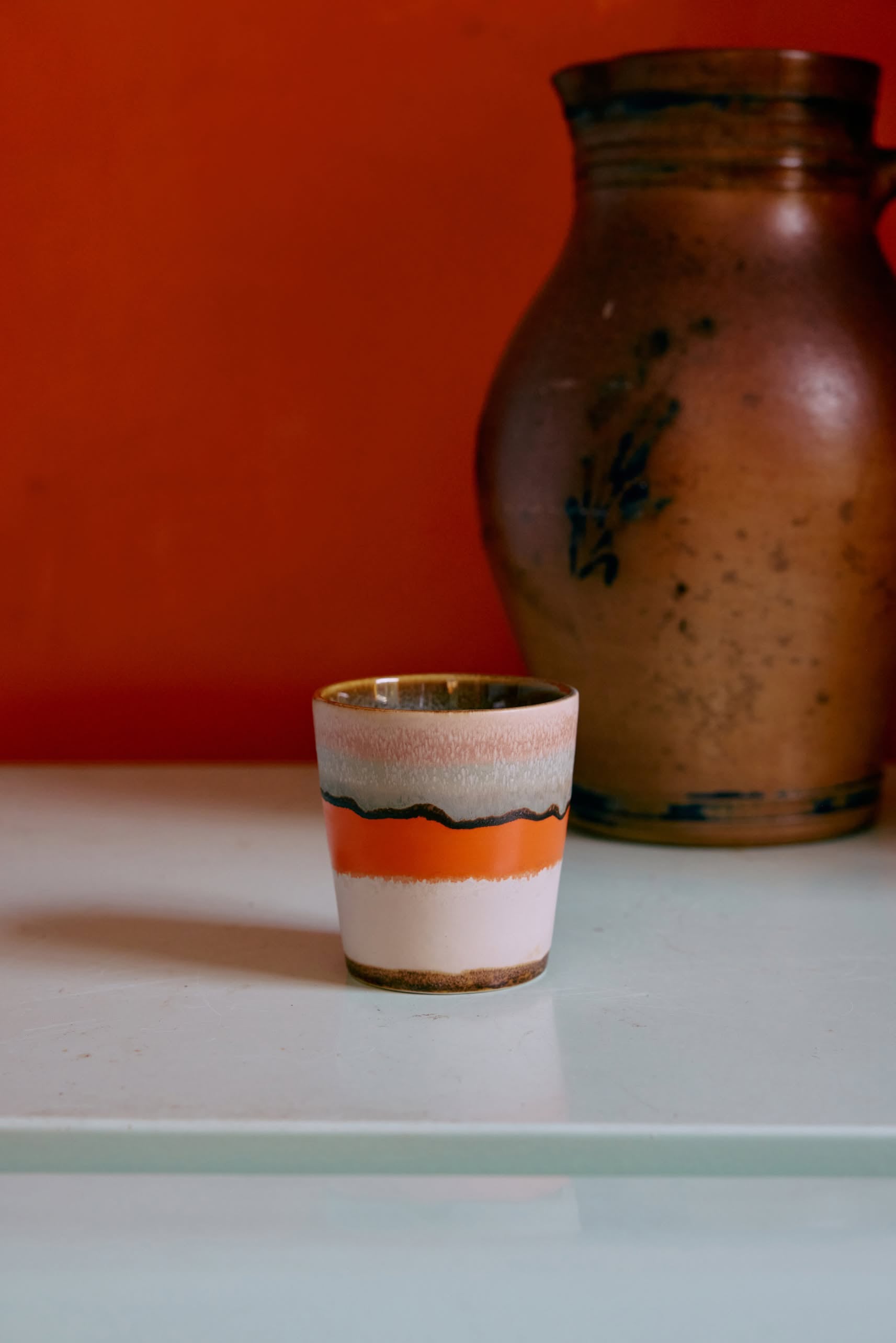 A HKLIVING 70s Ceramics: Coffee Mug with orange, white, and black stripes sits on a light surface, evoking retro design, while a large brown ceramic vase stands in the background against an orange wall. Set of 6 by HKLIVING.