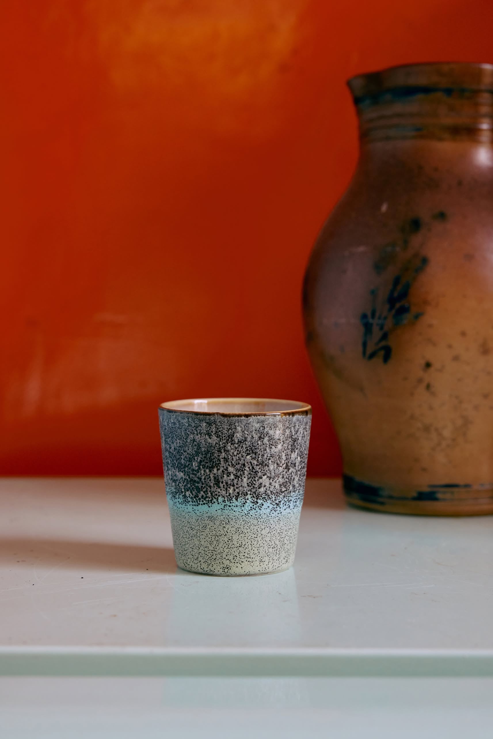 A set of 6 HKLIVING 70s Ceramics Coffee Mugs features textured, handcrafted glazing that fades from dark gray to light cream, capturing vintage style, all showcased on a pale surface with an orange wall backdrop.