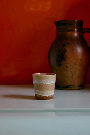 The HKLIVING 70s Ceramics Coffee Mug, from a set of 6 by HKLIVING, features a handcrafted striped glaze and sits on a light surface before a large brown earthenware jug with an orange wall in the background.