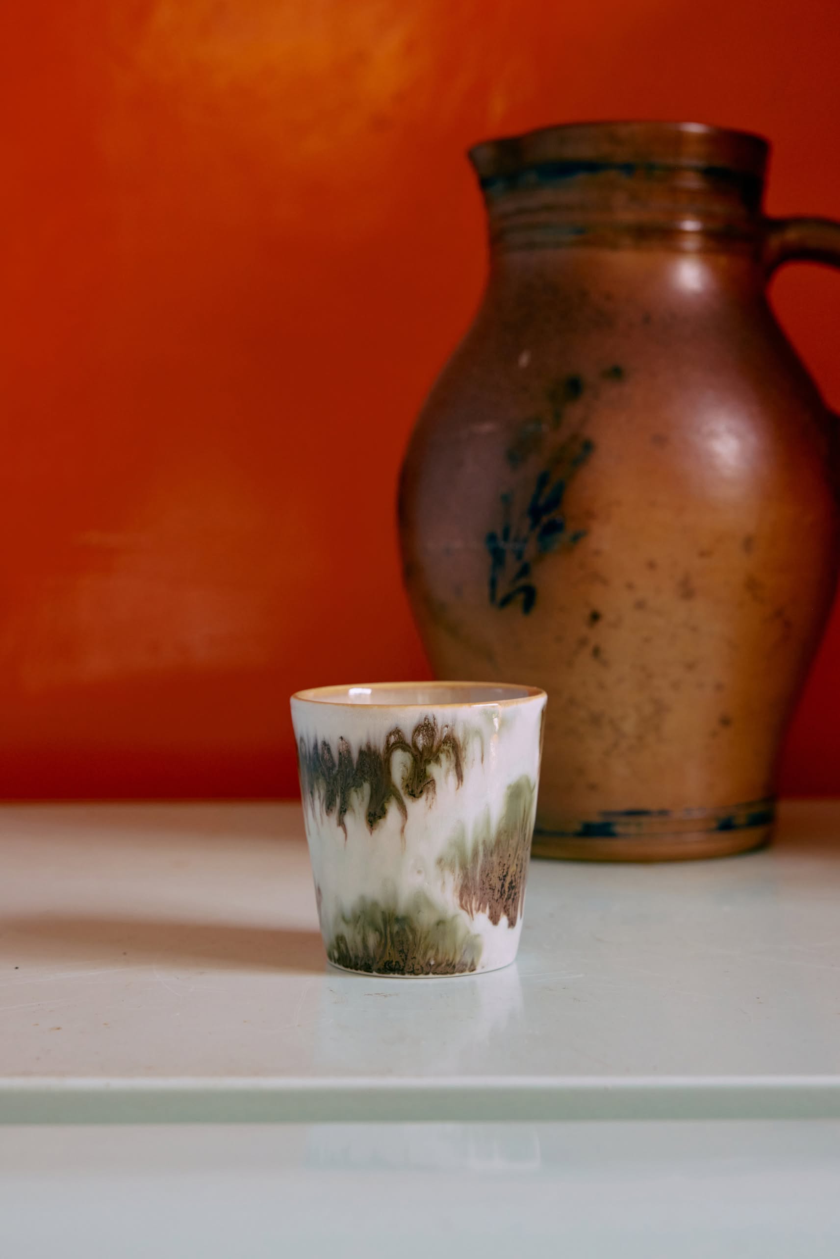 HKLIVING 70s Ceramics: Coffee Mugs, Set of 6 by HKLIVING are displayed on a white surface before a rustic jug, with a bright orange wall behind highlighting the vintage charm of these brown and green handcrafted mugs.