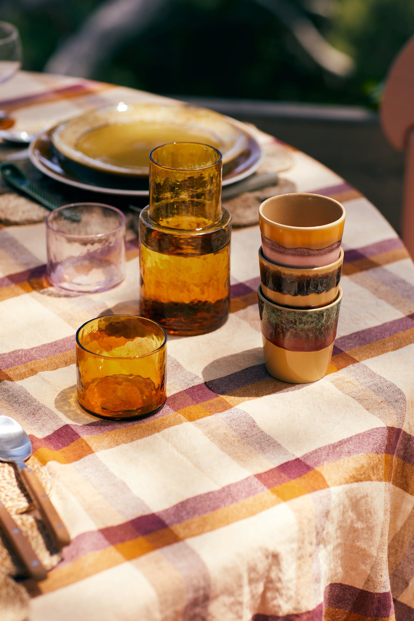 A close-up of a checkered tablecloth with stacked amber glasses and HKLIVING 70s Ceramics: Coffee Mugs, Set of 6, arranged outdoors in soft sunlight.