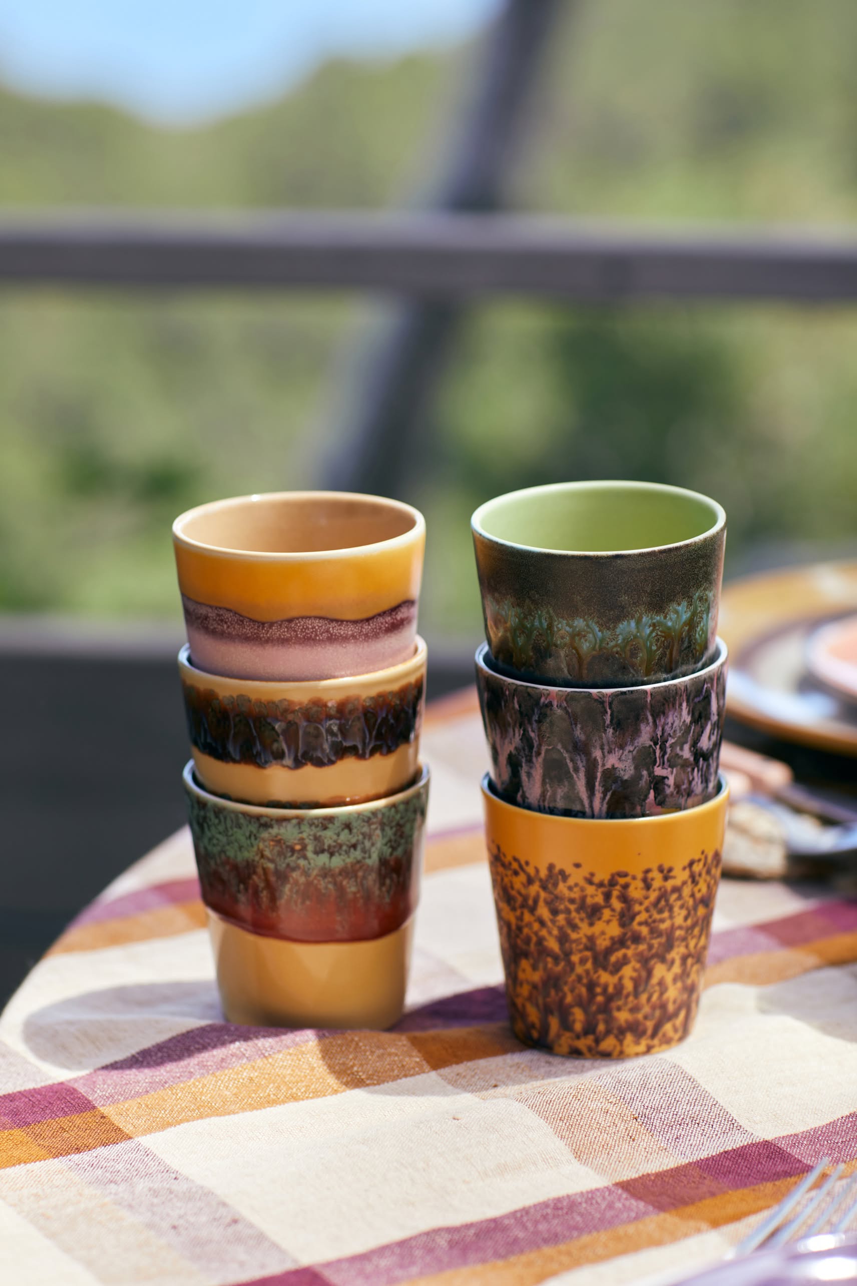 Six HKLIVING 70s Ceramics Coffee Mugs, Set of 6, are stacked in two piles outdoors on a checkered tablecloth with a blurred green background.