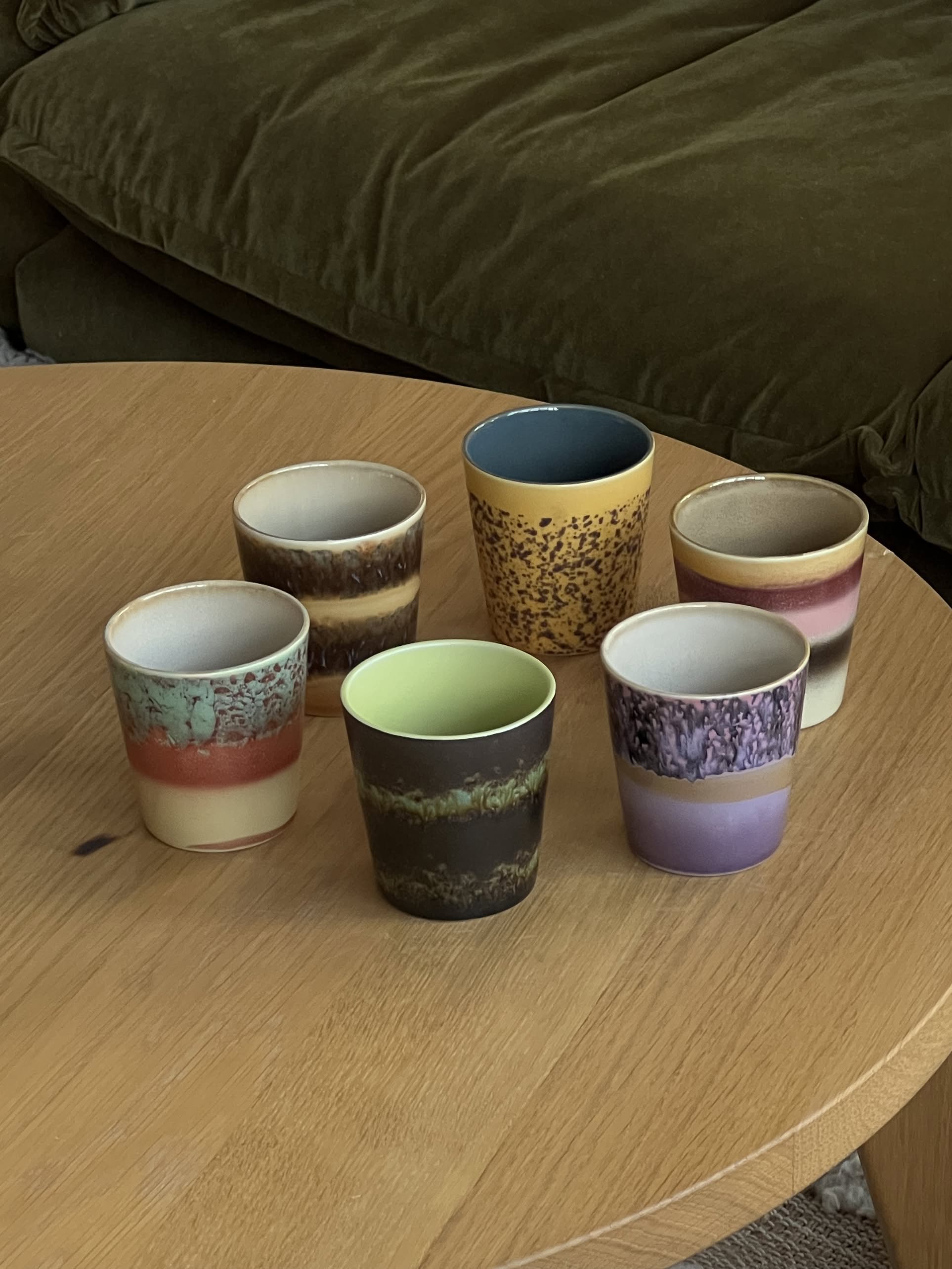 The HKLIVING 70s Ceramics: Coffee Mugs, Set of 6 features vibrant, handcrafted mugs arranged on a wooden table with a green sofa behind—channeling playful 1970s style by HKLIVING.