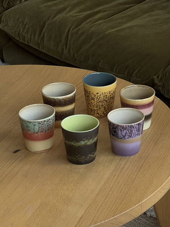 The HKLIVING 70s Ceramics: Coffee Mugs, Set of 6 features vibrant, handcrafted mugs arranged on a wooden table with a green sofa behind—channeling playful 1970s style by HKLIVING.