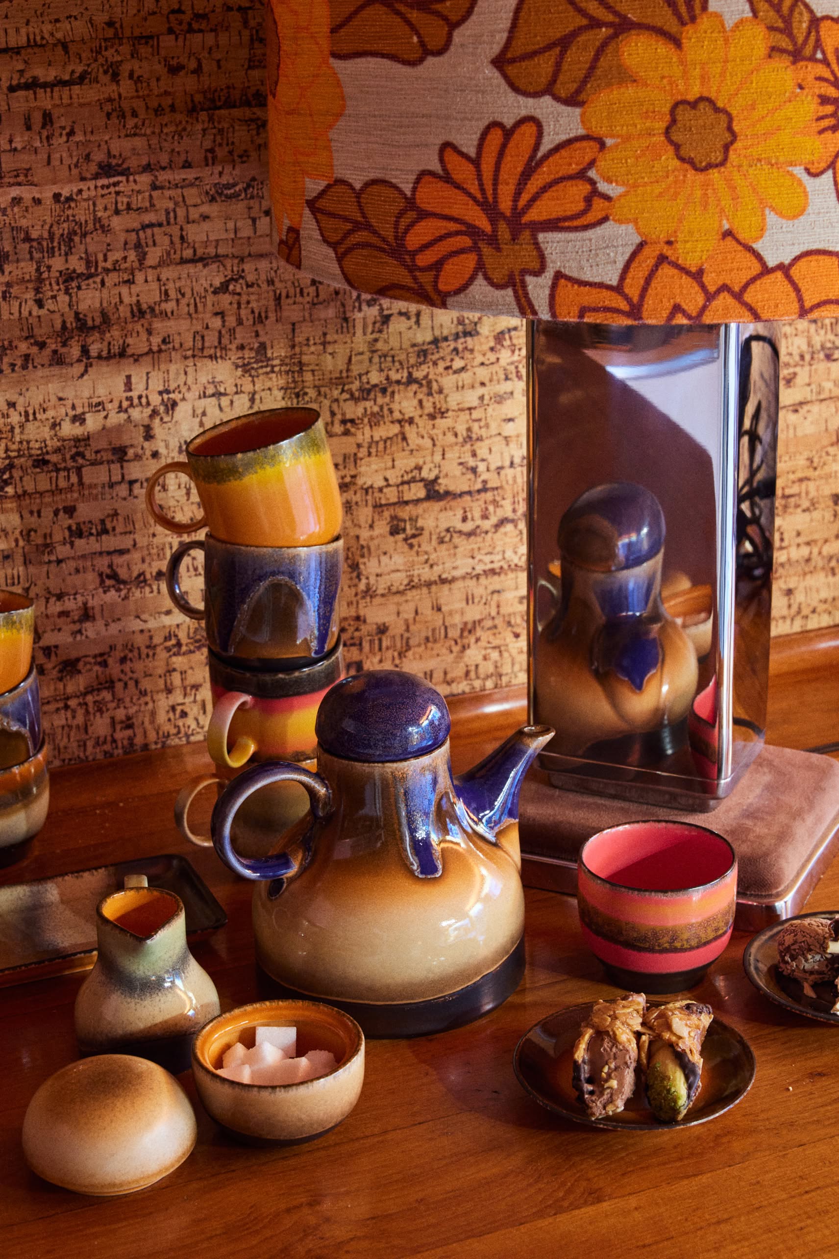 A retro table scene featuring the HKLIVING 70s Ceramics: Coffee Pot - Afternoon, mugs, sugar bowl, and cups arranged with pastries and a lamp with an orange floral shade on wood in earthy tones against a textured wall.