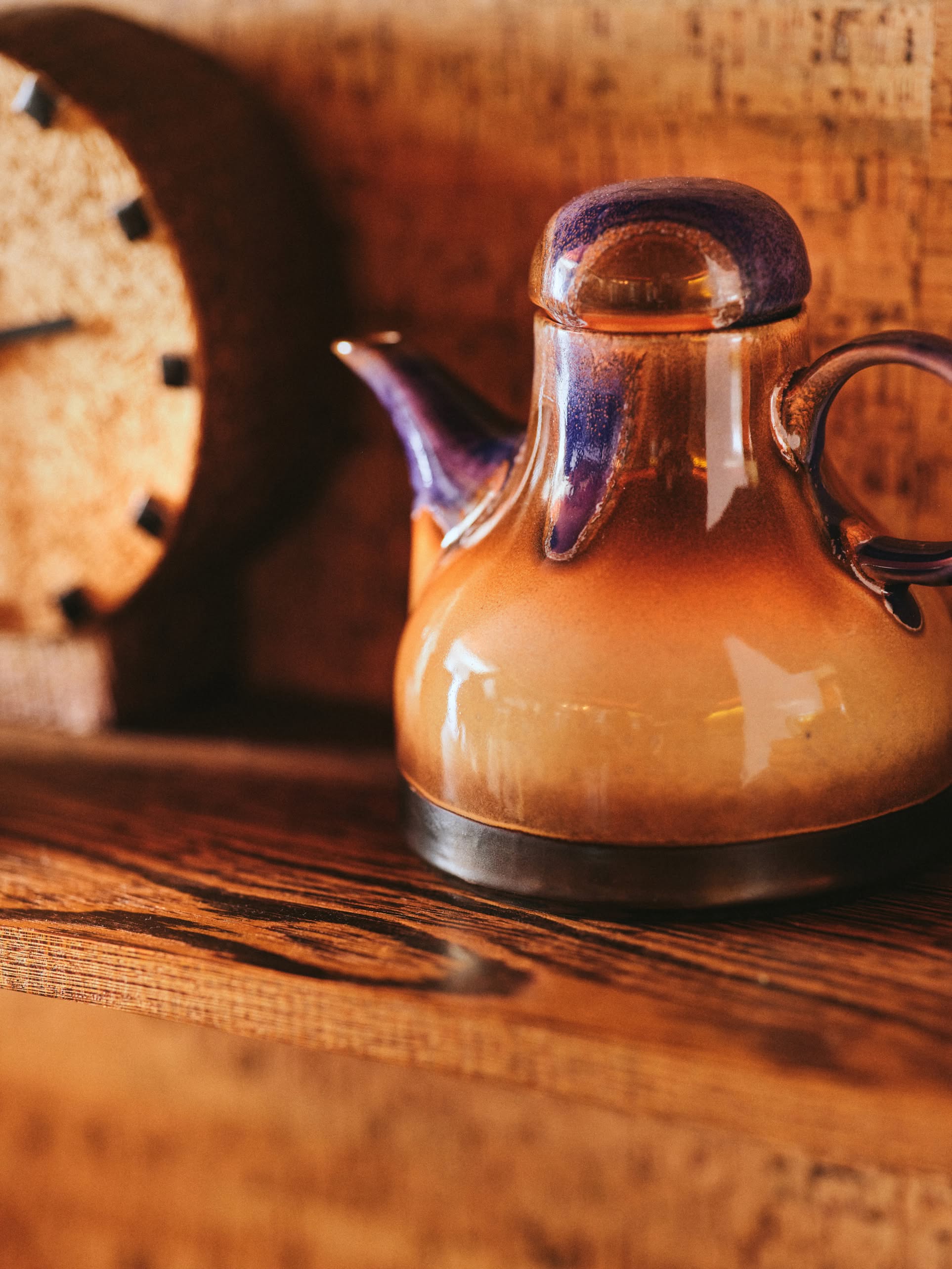 The HKLIVING 70s Ceramics: Coffee Pot - Afternoon, featuring an earthy brown and purple glaze, rests on a wooden shelf beside a blurred clock with black hour markers against a textured brown background.