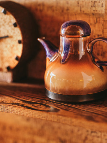 The HKLIVING 70s Ceramics: Coffee Pot - Afternoon, featuring an earthy brown and purple glaze, rests on a wooden shelf beside a blurred clock with black hour markers against a textured brown background.