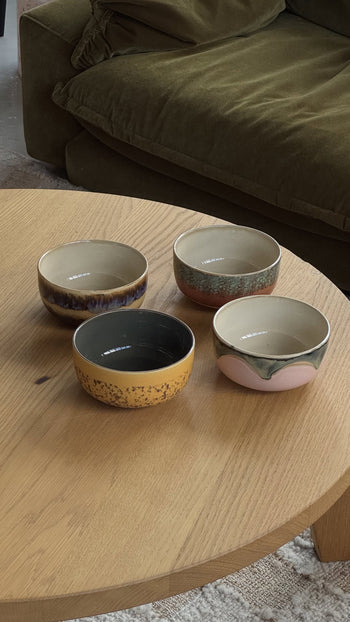 A set of 4 HKLIVING 70s Ceramics Dessert Bowls with colorful retro patterns is displayed in a square on a light wooden coffee table, positioned in front of a green couch.