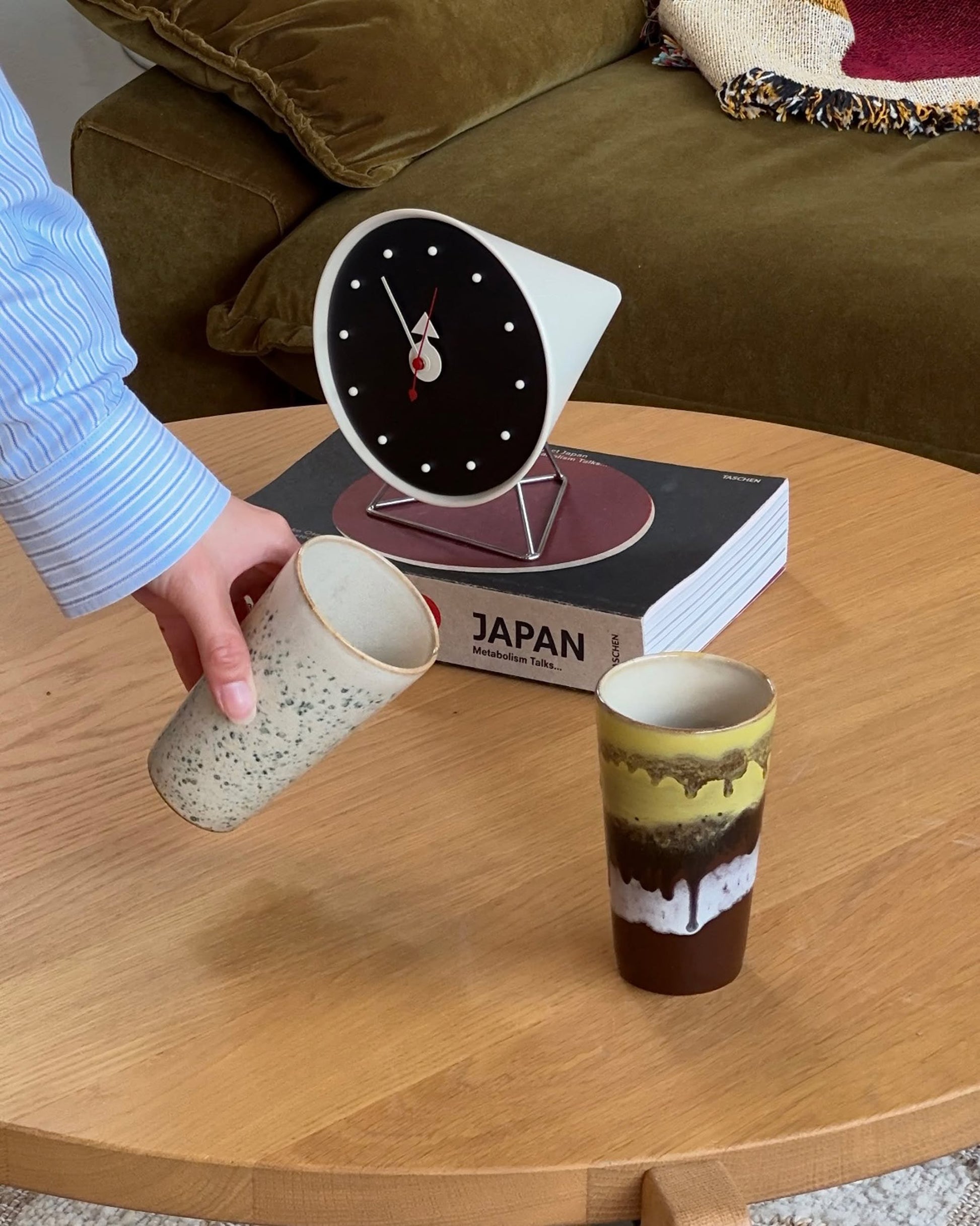 A hand holds an HKLIVING 70s Ceramics: Latte Mug above a wooden table with a matching mug, a clock, and the book "JAPAN"; a green couch is in the background.