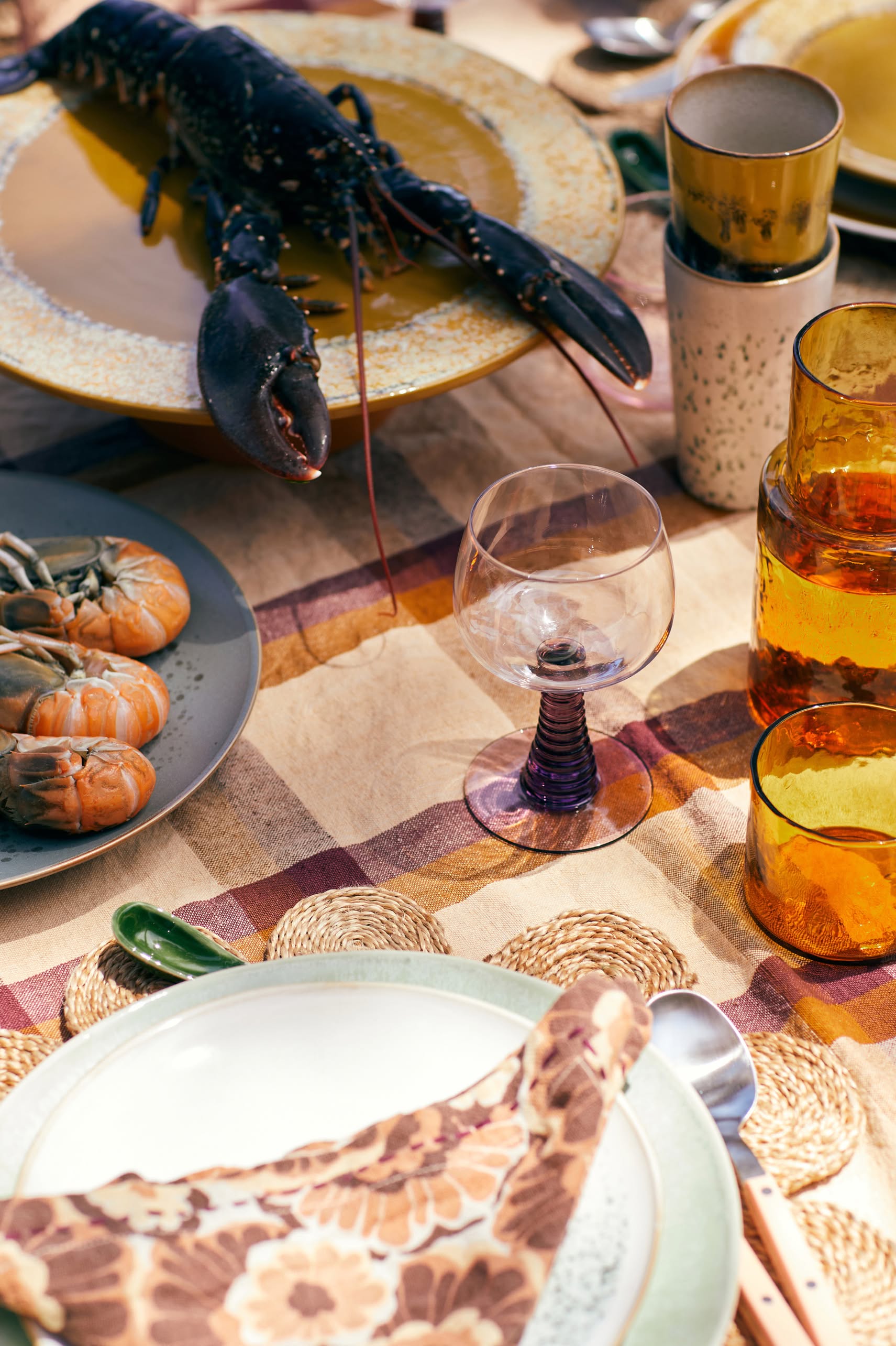 A close-up of a table set for a meal features a large lobster, prawns, amber glasses, an HKLIVING 70s Ceramics: Latte Mugs set, wine glass, patterned napkins, and woven placemats.