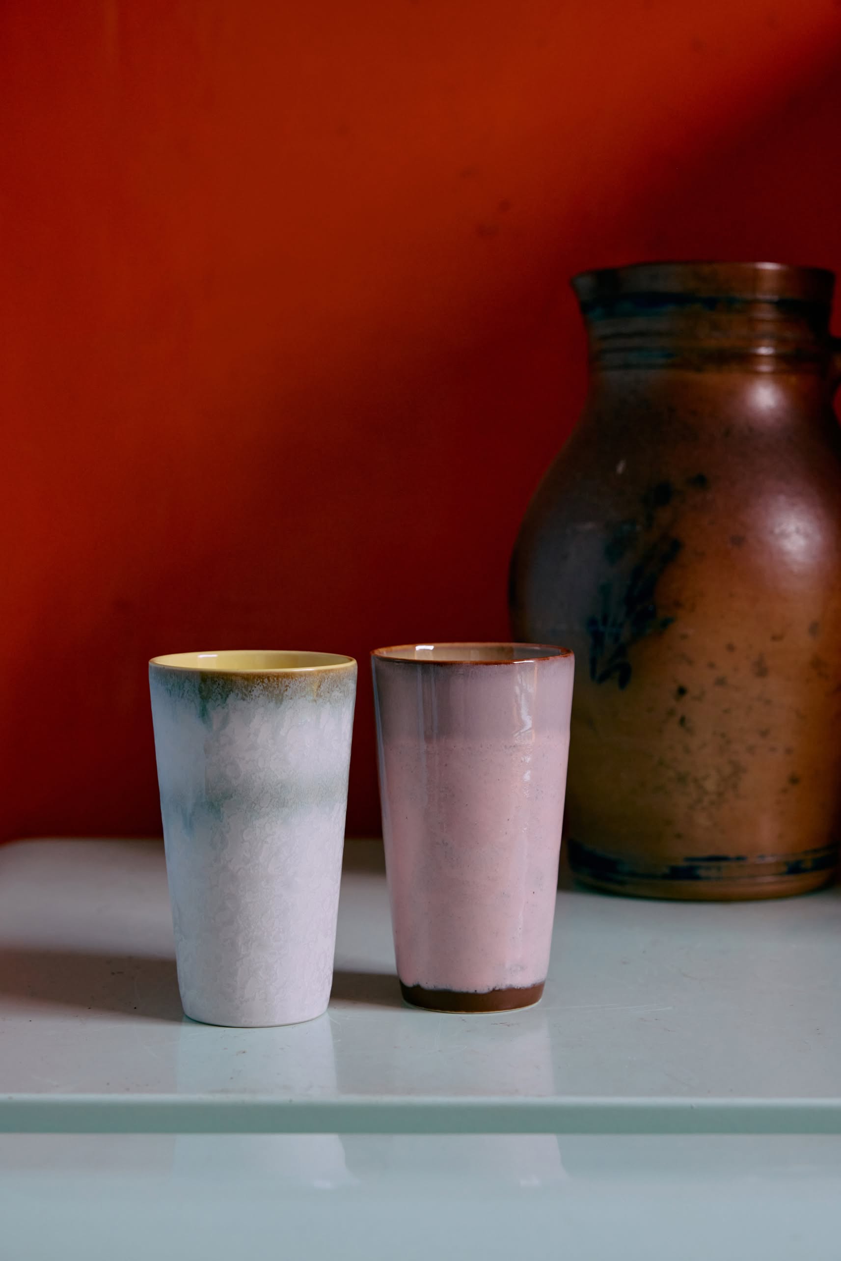 The HKLIVING 70s Ceramics: Latte Mugs, Set of 2, in white and light pink, are displayed side by side on a pale glossy surface with a brown vase behind them and an orange wall in the background.