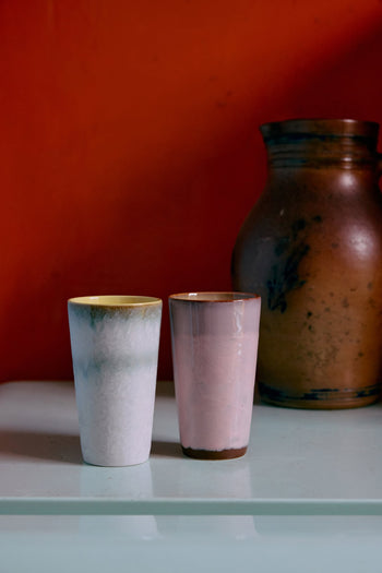 The HKLIVING 70s Ceramics: Latte Mugs, Set of 2, in white and light pink, are displayed side by side on a pale glossy surface with a brown vase behind them and an orange wall in the background.