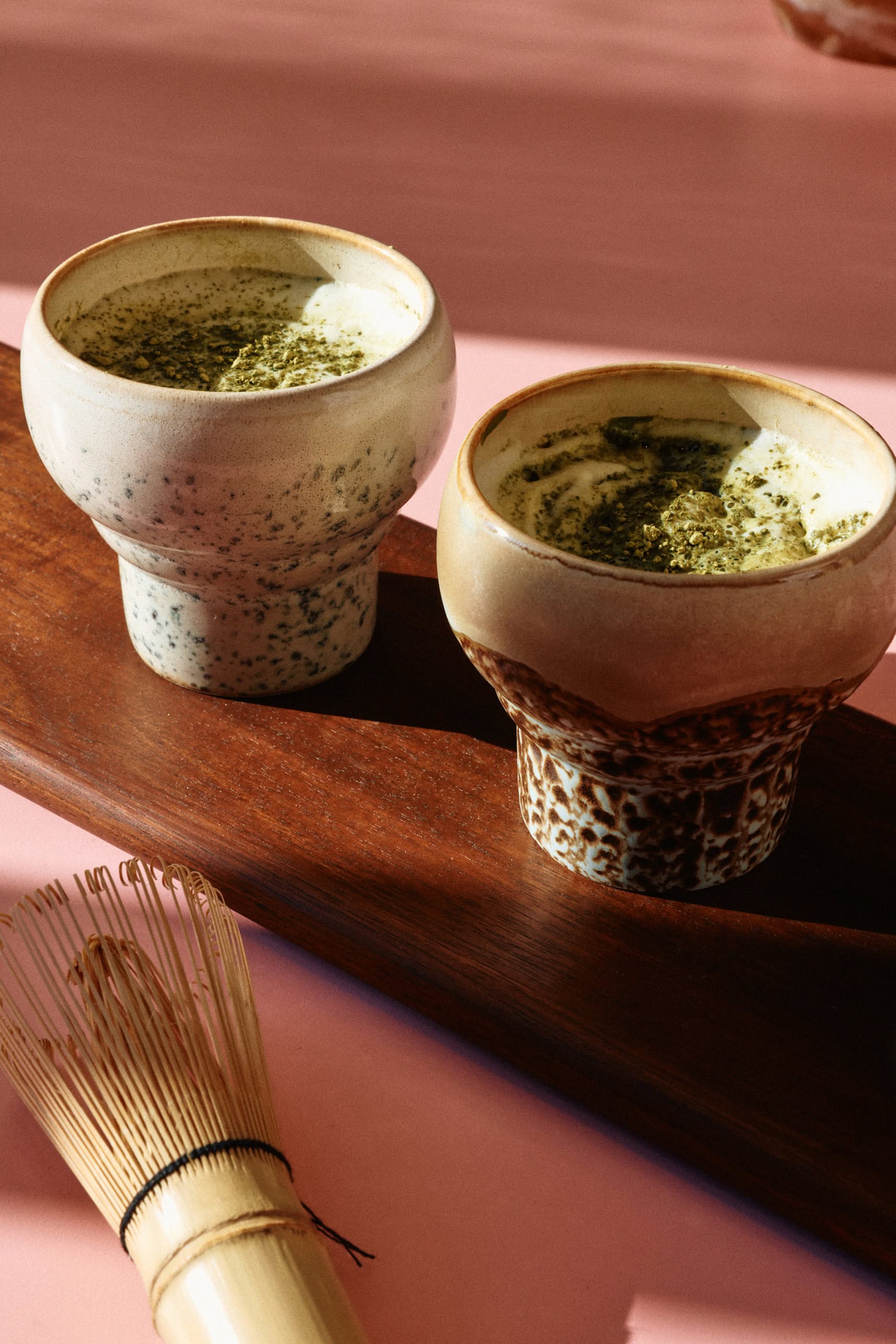 Two HKLIVING 70s Ceramics Lungo Mugs, Set of 2, are filled with frothy matcha tea on a wooden tray with a bamboo whisk nearby atop a pink surface, all illuminated by warm sunlight.
