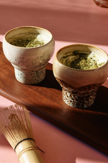 Two HKLIVING 70s Ceramics Lungo Mugs, Set of 2, are filled with frothy matcha tea on a wooden tray with a bamboo whisk nearby atop a pink surface, all illuminated by warm sunlight.