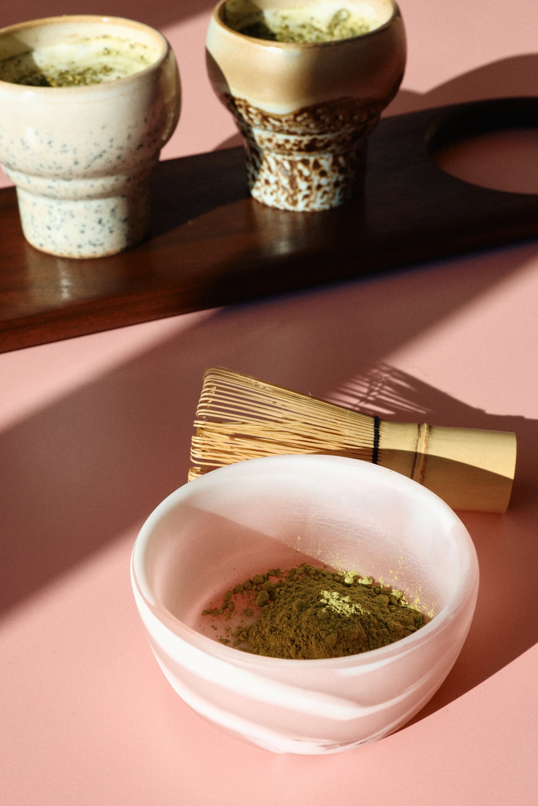 A ceramic bowl with matcha powder and a bamboo whisk sit on a pink surface, while two HKLIVING 70s Ceramics: Lungo Mugs (Set of 2) rest on a wooden tray in the background. Sunlight casts soft shadows across the scene.