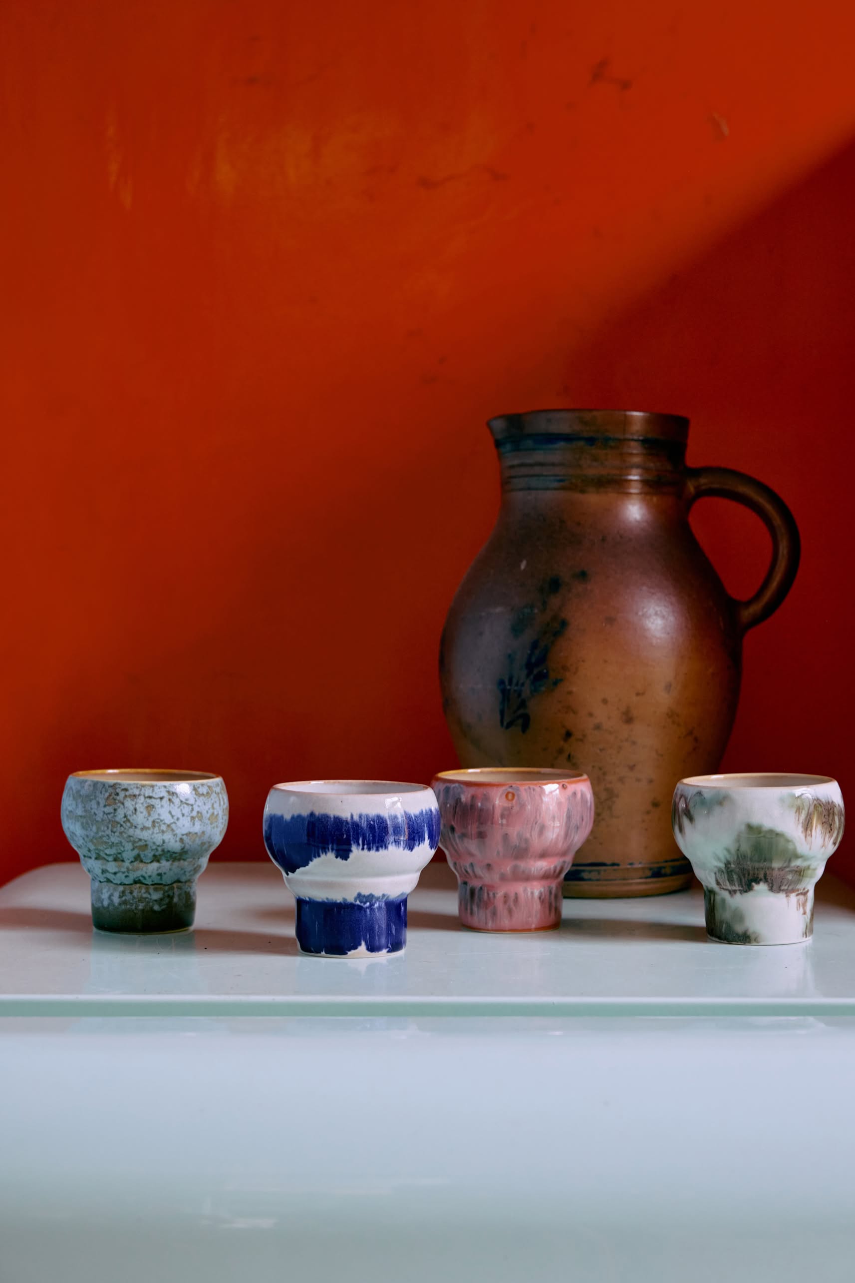 Two HKLIVING 70s Ceramics Lungo Mugs are displayed on a white surface before a large brown jug with handle, set against a vibrant orange-red background.