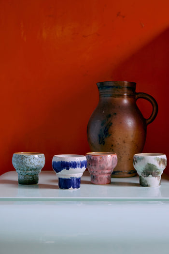 Two HKLIVING 70s Ceramics Lungo Mugs are displayed on a white surface before a large brown jug with handle, set against a vibrant orange-red background.