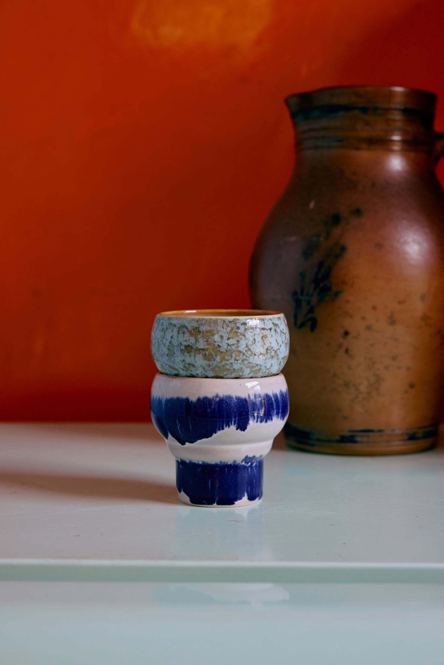 The HKLIVING 70s Ceramics: Lungo Mugs, Set of 2 features small, two-toned blue and white mugs with a textured light blue top, placed on white surfaces; a large brown jug stands out against an orange backdrop.