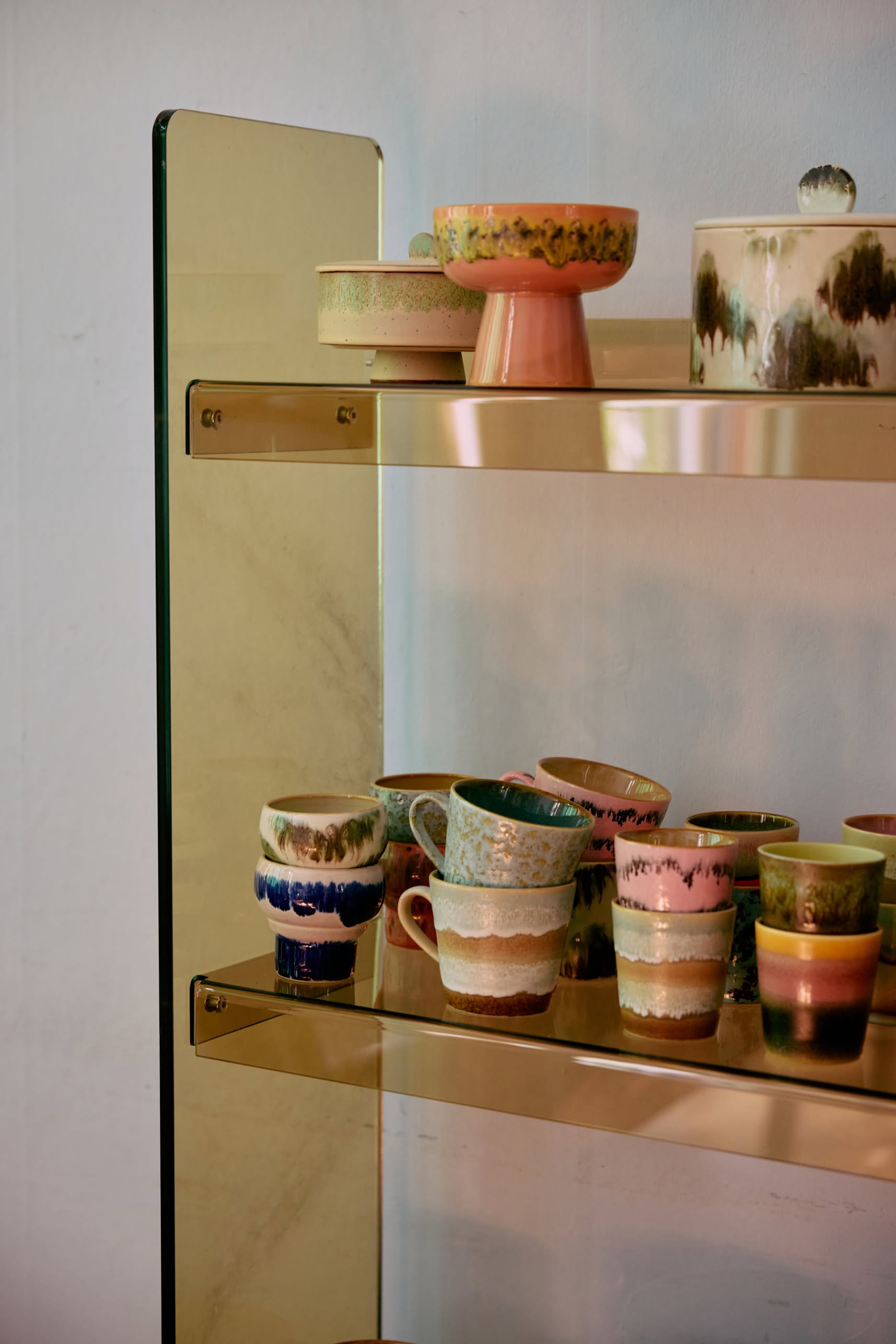 A gold-framed glass shelf displays HKLIVING 70s Ceramics Lungo Mugs (Set of 2) alongside retro ceramic cups and bowls in pastel and earthy tones, all with textured, glazed finishes, against a light-colored wall.