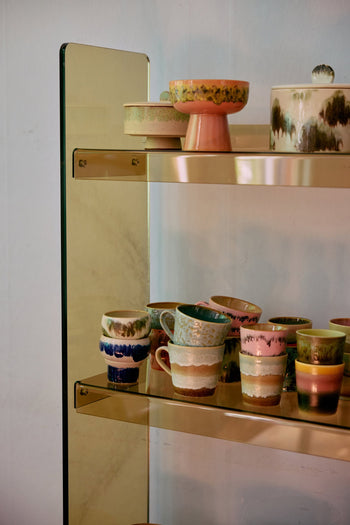 A gold-framed glass shelf displays HKLIVING 70s Ceramics Lungo Mugs (Set of 2) alongside retro ceramic cups and bowls in pastel and earthy tones, all with textured, glazed finishes, against a light-colored wall.