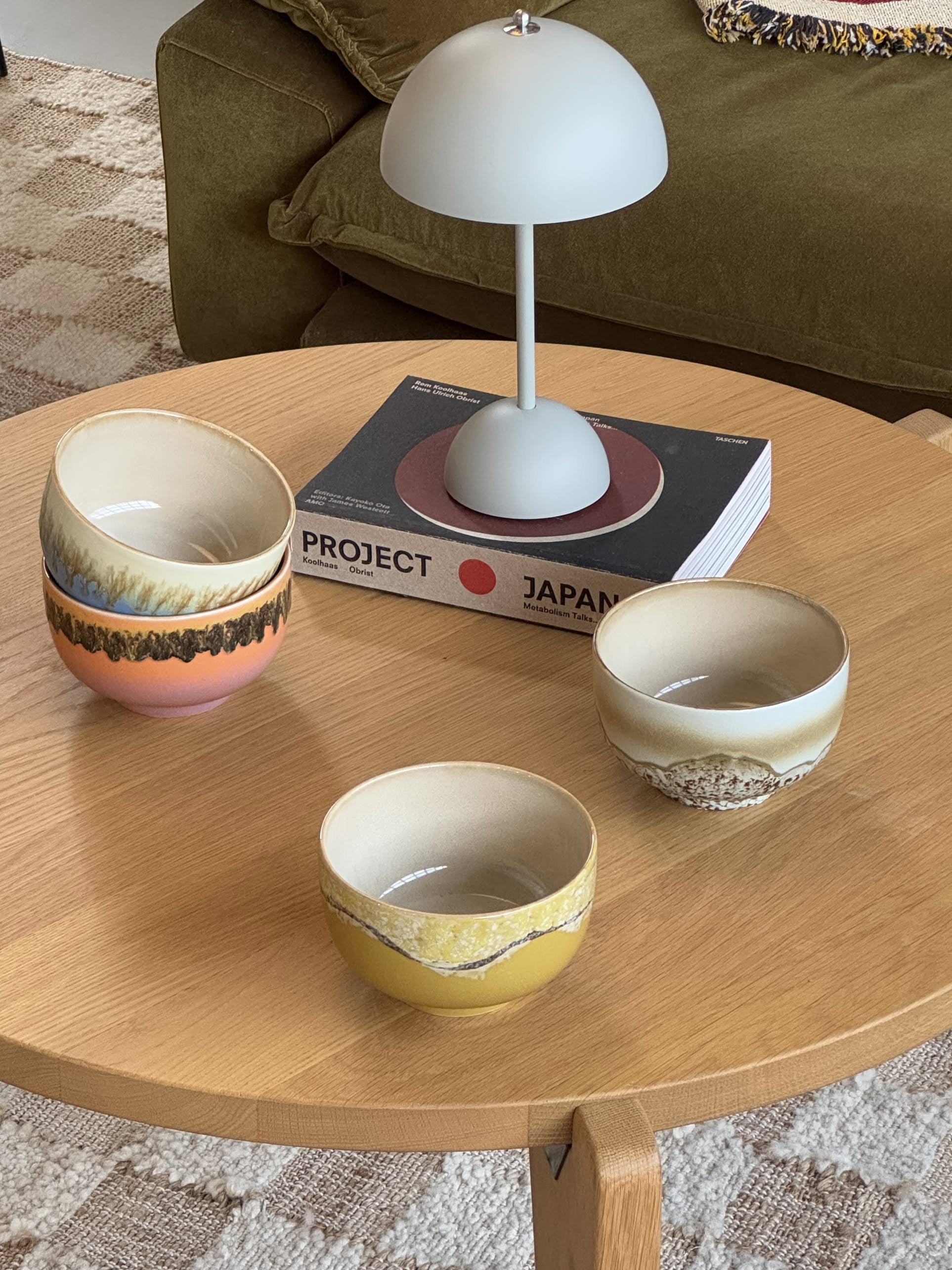 Four HKLIVING 70s Ceramics Noodle Bowls sit on a round wooden table beside a modern lamp and the book PROJECT JAPAN, with a green sofa and patterned rug adding to the retro-inspired vibe.