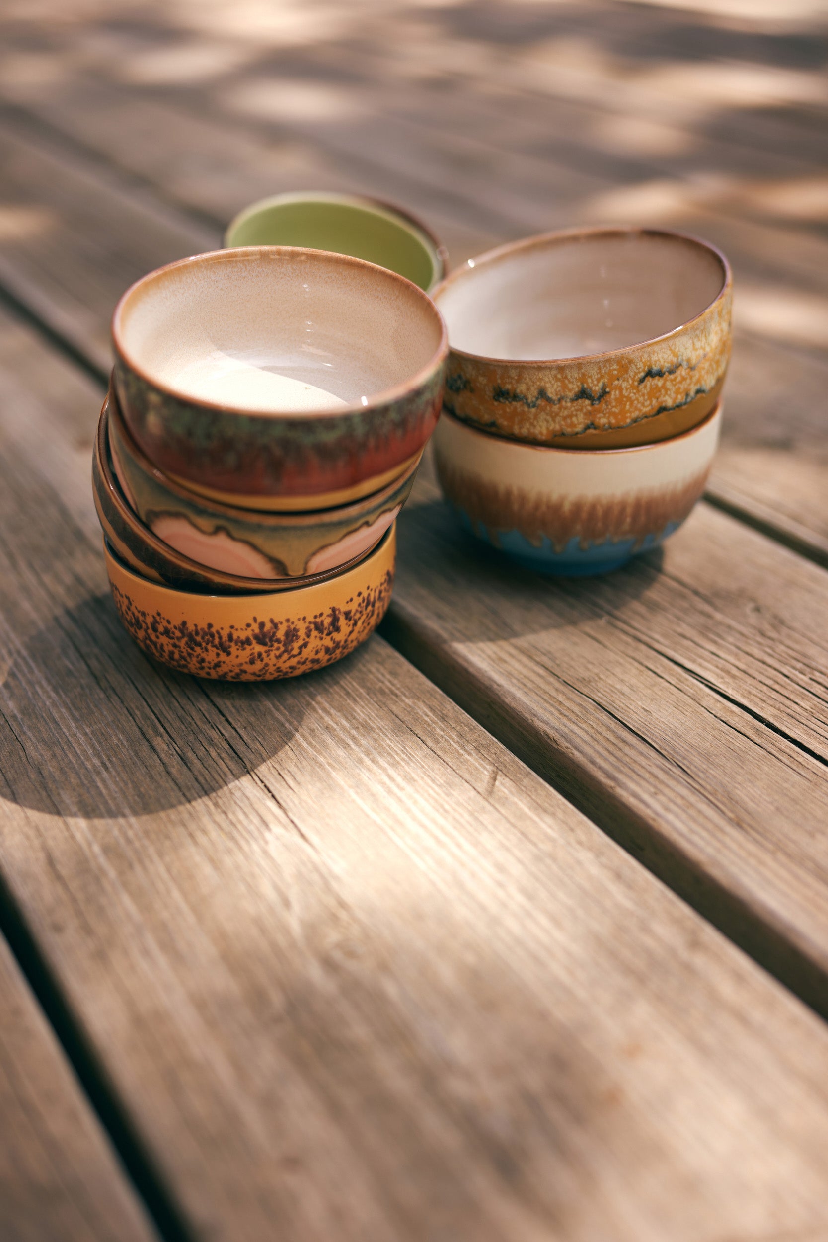 Four HKLIVING 70s Ceramics Noodle Bowls, each featuring unique earthy patterns and colors, are stacked on a sunlit wooden surface, casting soft shadows.