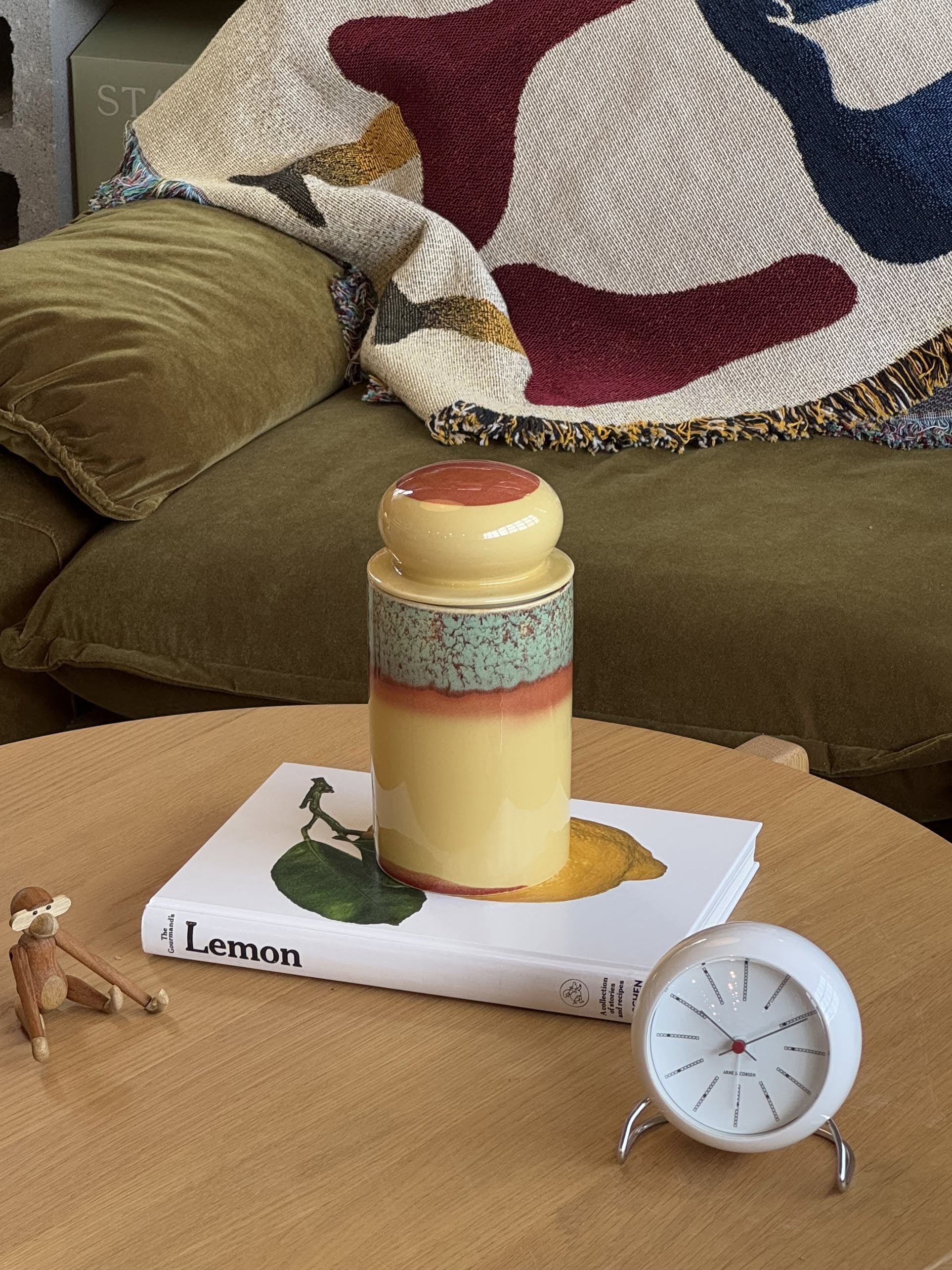 A cozy living room features a green velvet couch, a colorful blanket, and on the wooden table, the HKLIVING 70s Ceramics: Storage Jar - Cove, a book titled Lemon, a small white clock, and a wooden monkey figurine.