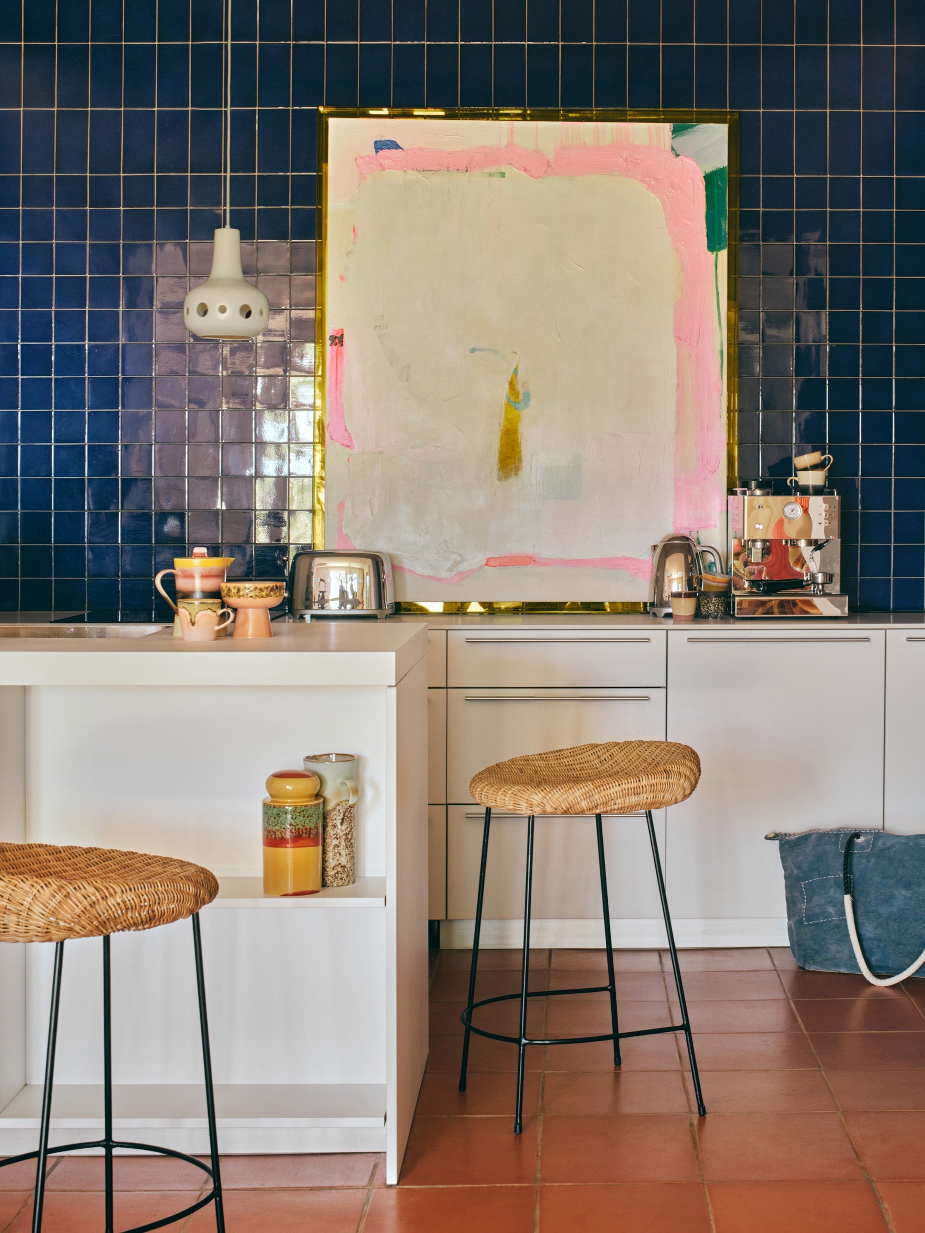 A modern kitchen with dark blue tiled walls, white cabinets, rattan bar stools, and a large abstract painting. On the counter are a coffee machine, toasters, mugs, and the HKLIVING 70s Ceramics: Storage Jar - Cove by HKLIVING; a woven bag sits on the floor.