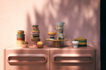 A vibrant display of HKLIVING 70s Ceramics, featuring the Storage Jar - Cove along with cups, bowls, and a pitcher on a pink cabinet against a sunlit pink wall adorned with leafy shadows.