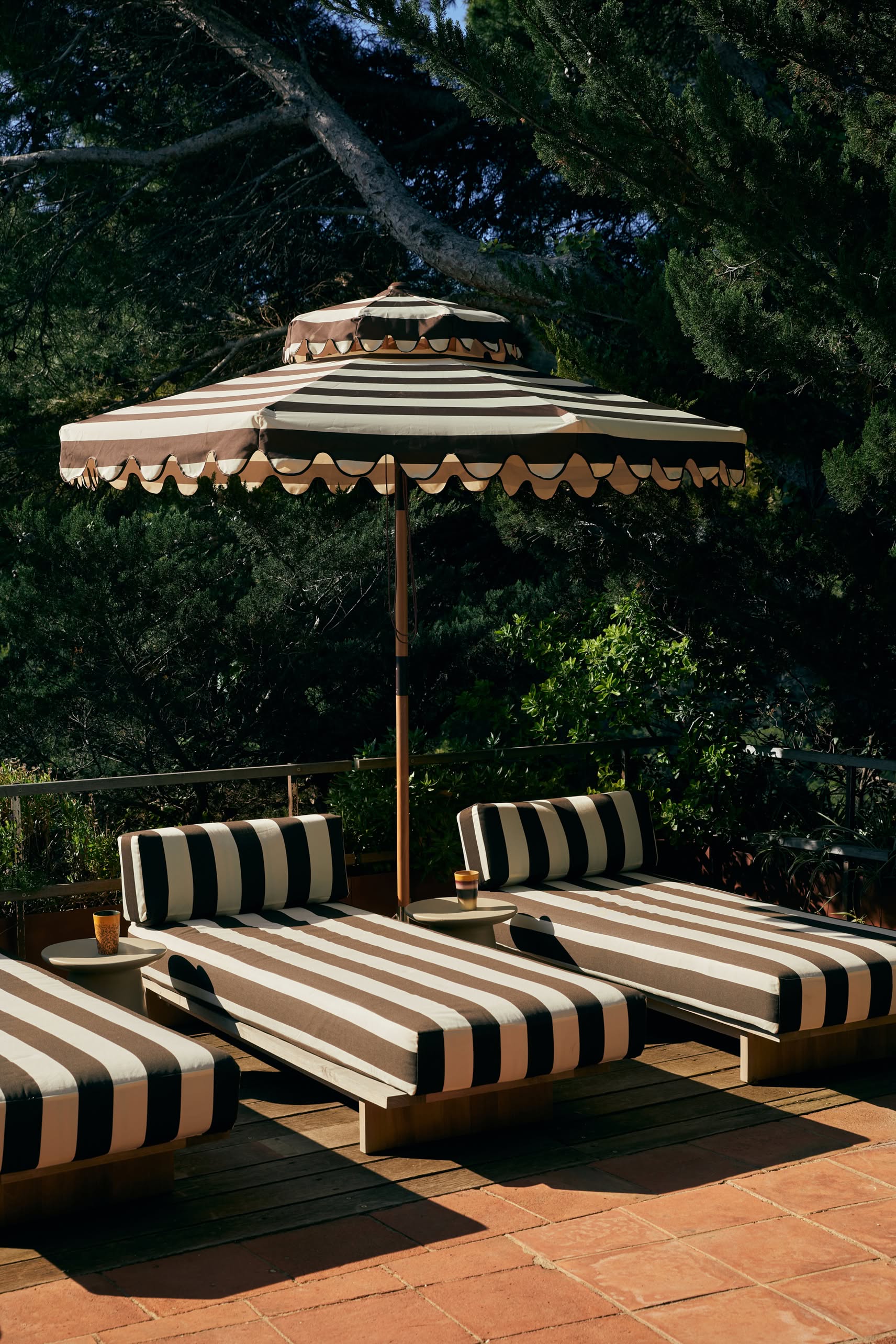 Four black-and-white striped lounge chairs sit under a matching umbrella on a terracotta patio with lush greenery. A set of HKLIVING 70s Ceramics Tea Mugs adds vintage charm to the serene outdoor setting.