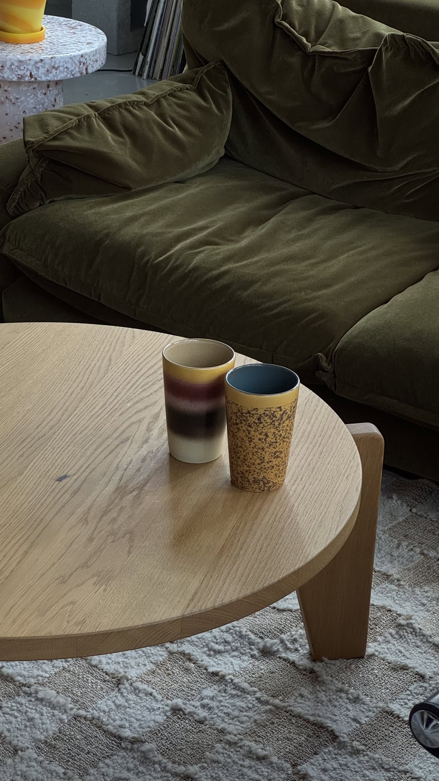 The HKLIVING 70s Ceramics: Tea Mugs, Set of 2 by HKLIVING—a brown and cream striped mug and a yellow speckled mug—sit on a round wooden coffee table in front of a green velvet sofa.