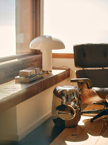 A cozy reading nook with a modern lounge chair, the HKLIVING Currents Chrome Side Table by HKLIVING, books, a white mushroom-shaped lamp, and sunlight pouring through a window—an ideal setup for contemporary interiors.
