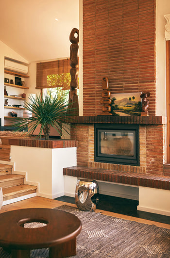 A cozy living room with a brick fireplace, modern sculptures, a potted plant, and a wooden coffee table. Contemporary style stands out with the HKLIVING Currents Chrome Side Table, patterned rug, bookshelves with decor, and sunlight streaming in.