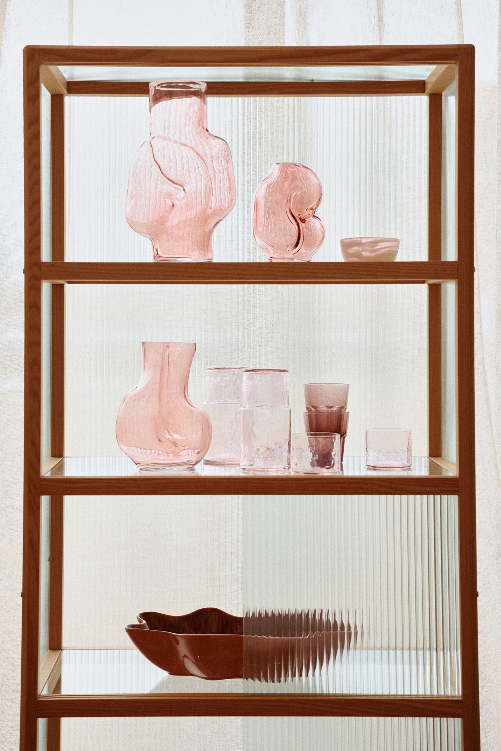 A wooden and glass shelf displays HKLIVING Gleam Glass Vases in pink, clear, and olive green, each with unique artistic shapes. Sunlight filters through sheer curtains behind them.