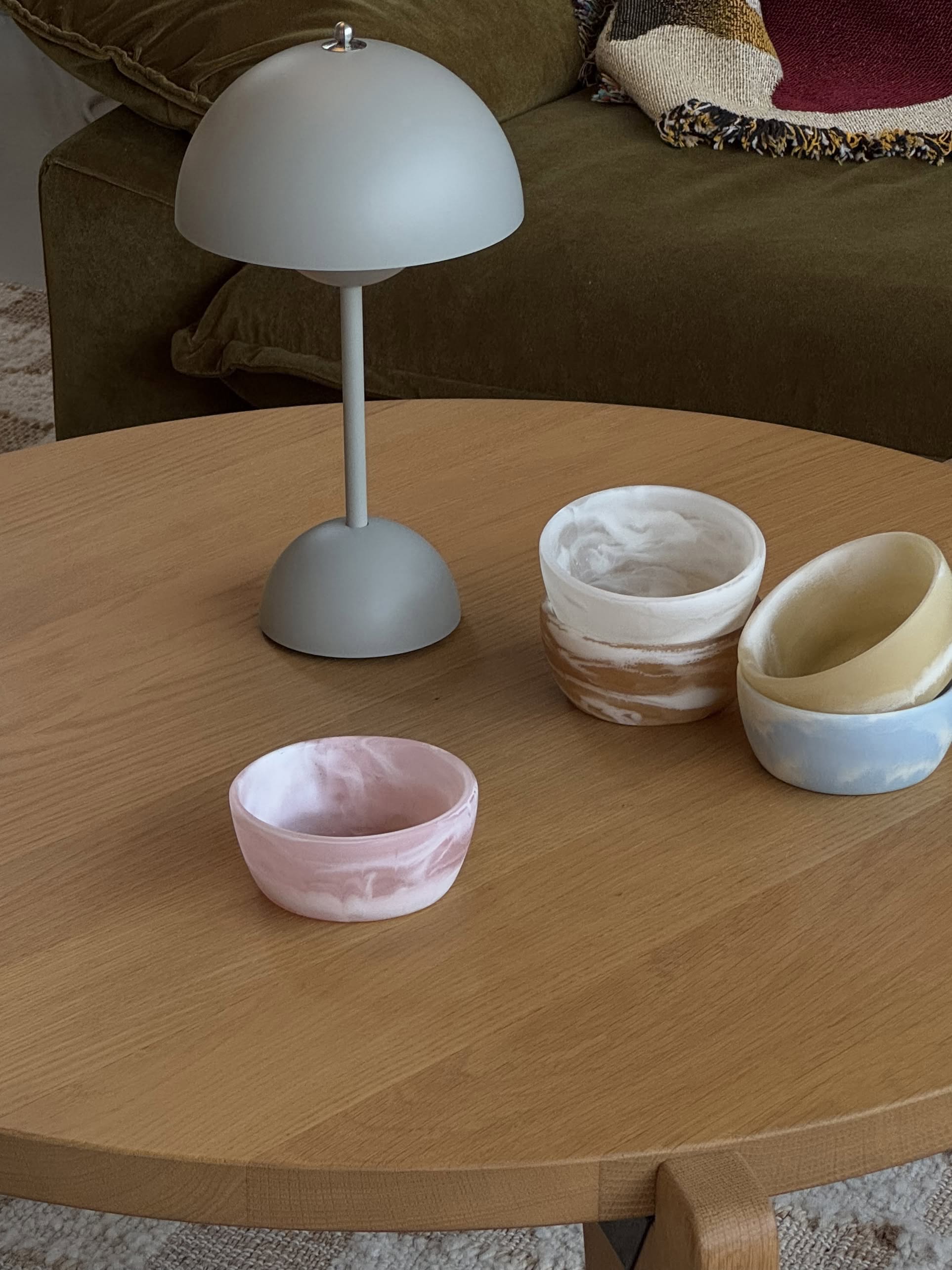 A round wooden coffee table holds a modern gray lamp and four HKLIVING Patio Bowls by HKLIVING in pink, brown, white, and blue marbled resin. A green sofa and a woven cushion finish this stylish setup—ideal for outdoor entertaining.